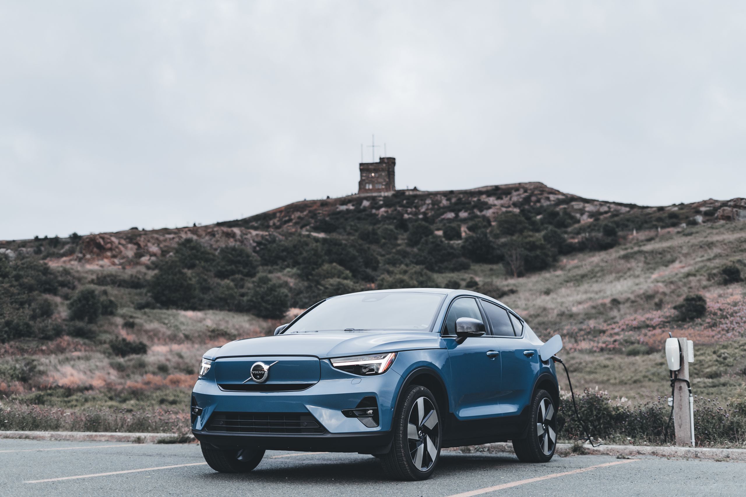 A blue Volvo electric car charging in a scenic outdoor location with a hill in the background.